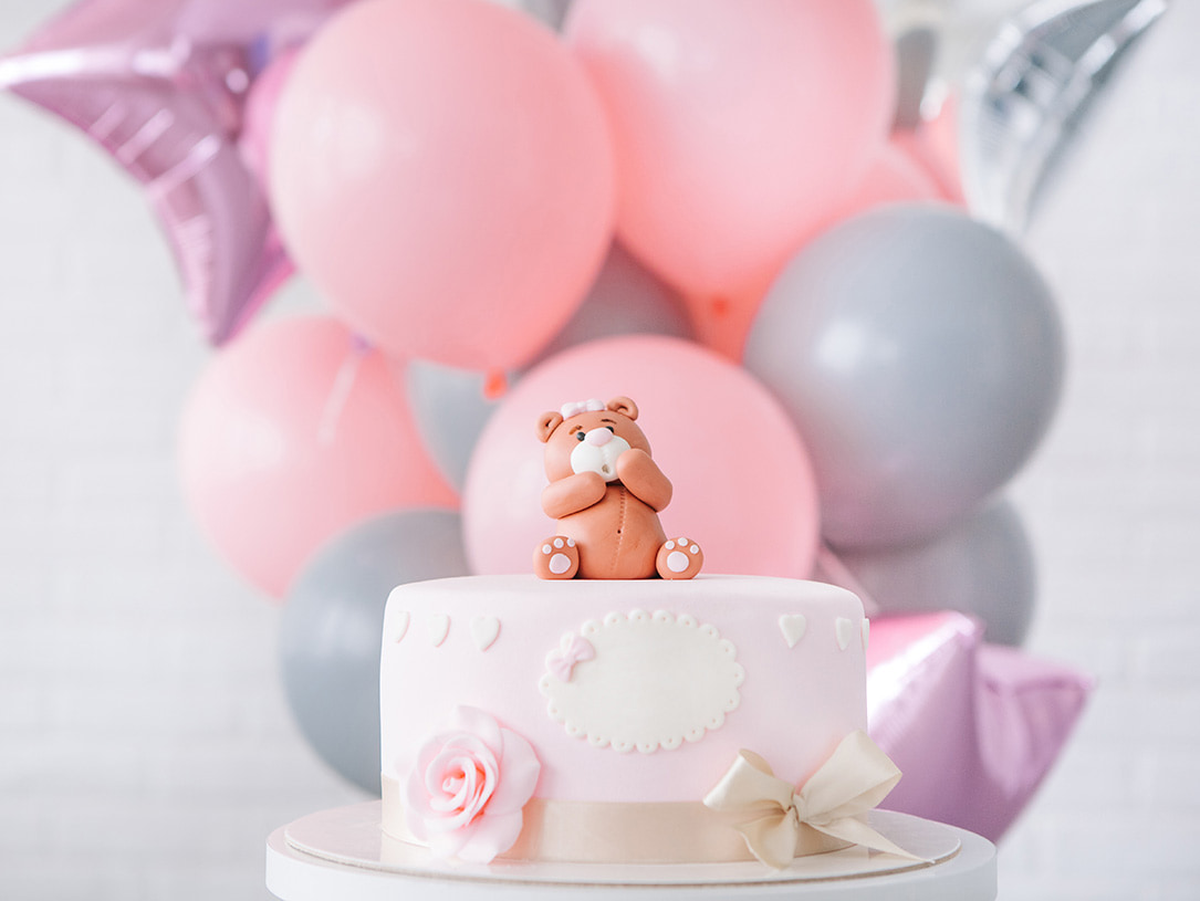 Mit einem kleinen Bären verzierte rosafarbene Torte mit Blümchen und Schleife dekoriert und Luftballons im Hintergrund