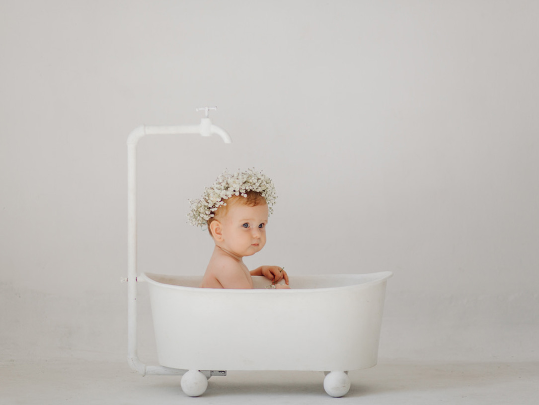 Baby mit Blumenkranz auf dem Kopf sitzt in einer kleinen freistehenden Badewanne