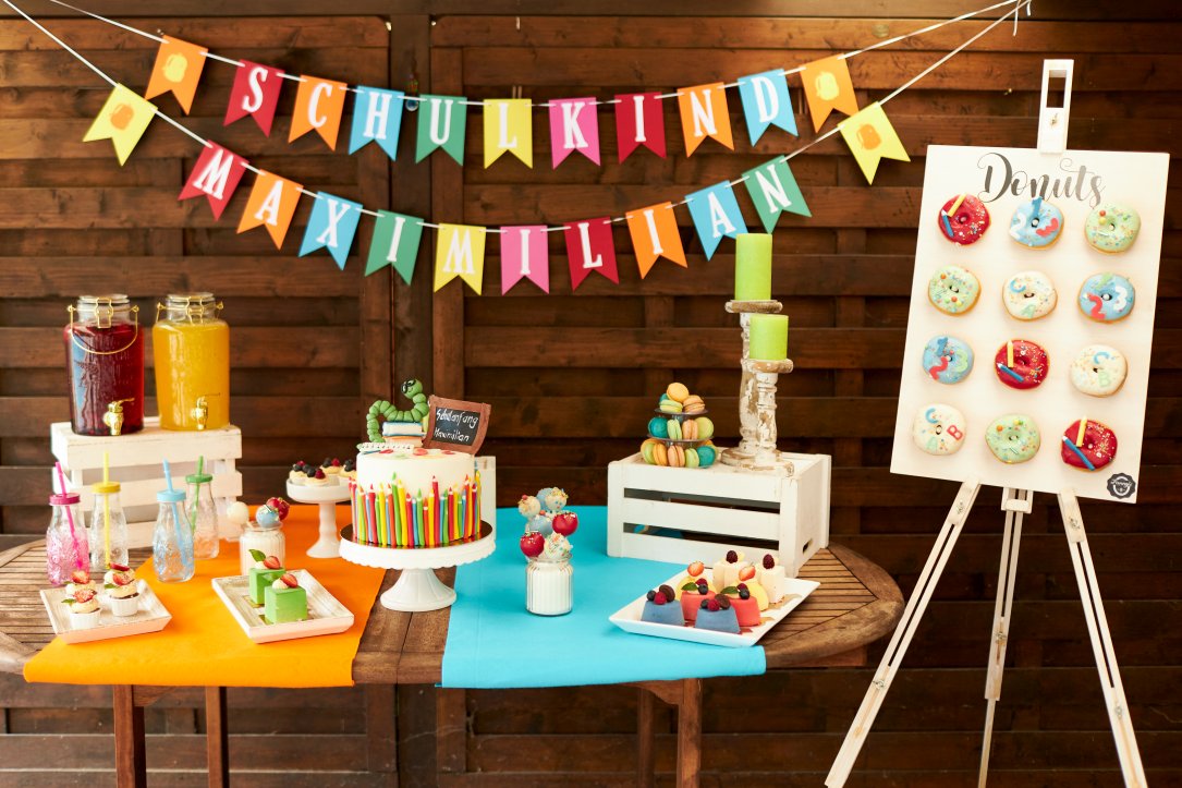 Bunt dekorierter Geburtstagstisch mit Torte und anderen Leckereien sowie einer Donuts-Tafel und einer Wimpelkette im Hintergrund