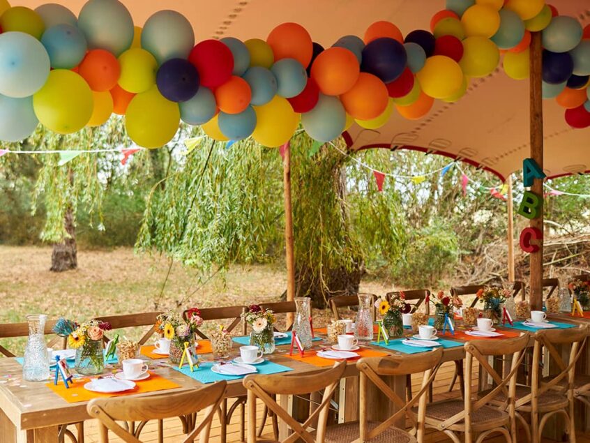 Bunt geschmückte Tafel mit Holzmöbeln und Zelt mit vielen bunten Luftballons im Freien