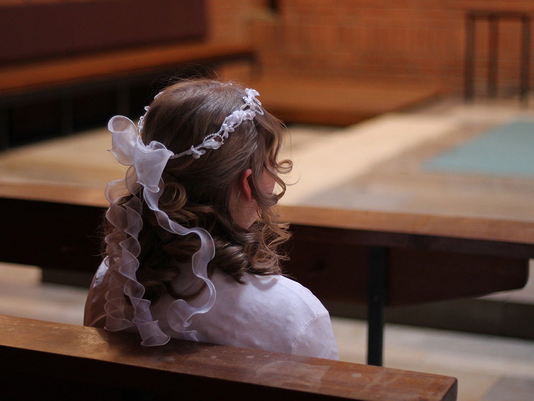 Kleines Mädchen sitzt auf Kirchengestühl und trägt ein weißes Oberteil und weißen Haarschmuck