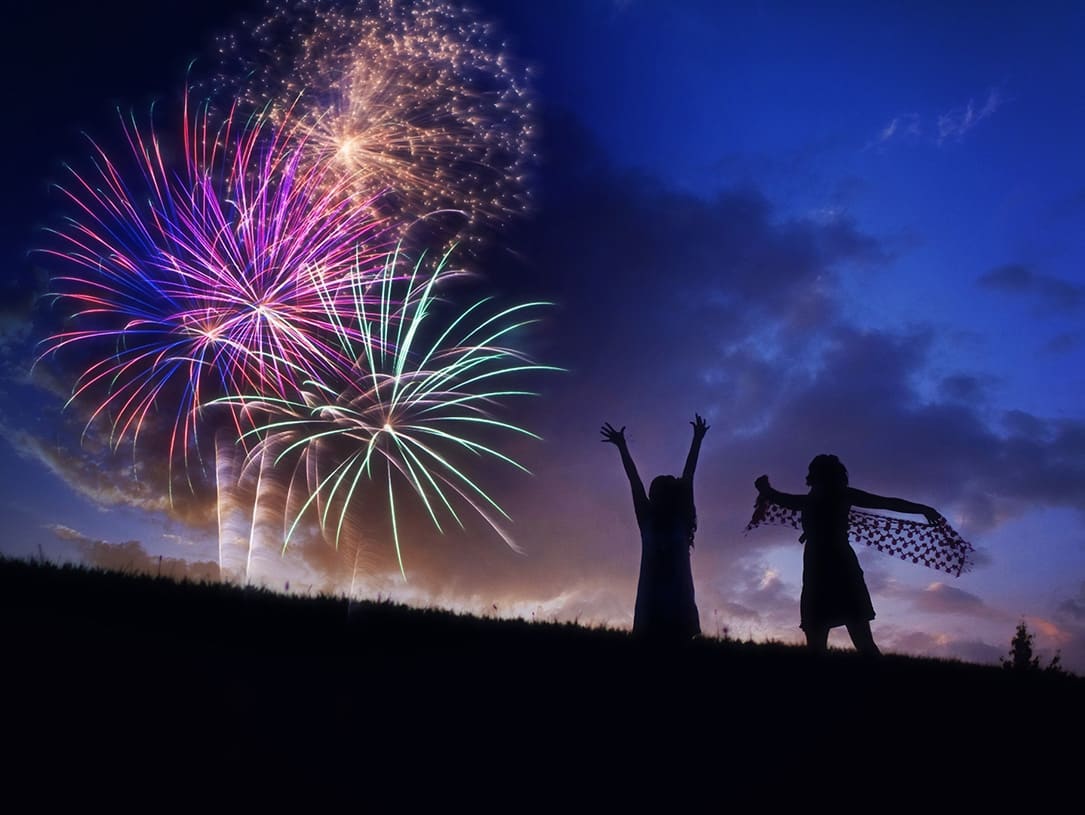 Zwei Personen stehen nachts auf einer Wiese und freuen sich über buntes Feuerwerk im Hintergrund
