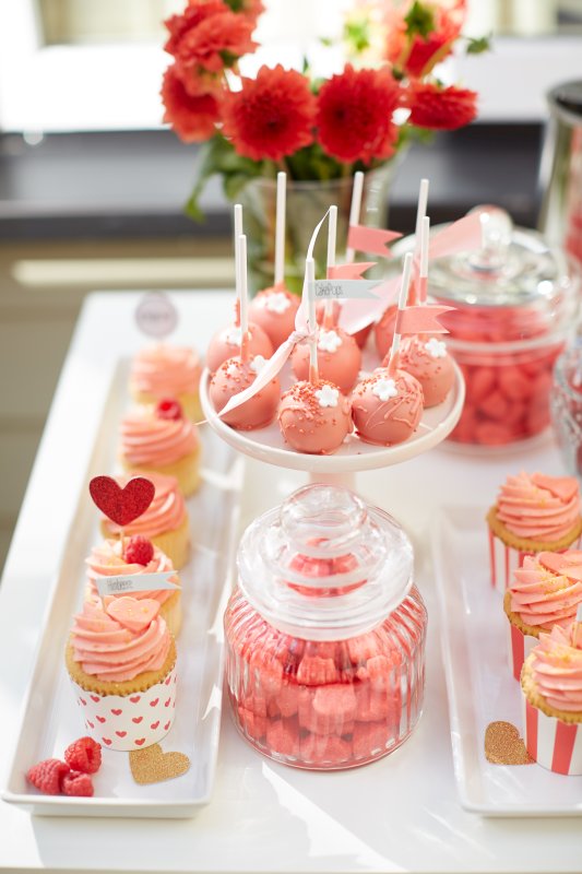 Rosa gehaltene Muffings, Cakepops und andere Leckereien auf einem Tisch mit roten Blumen im Hintergrund