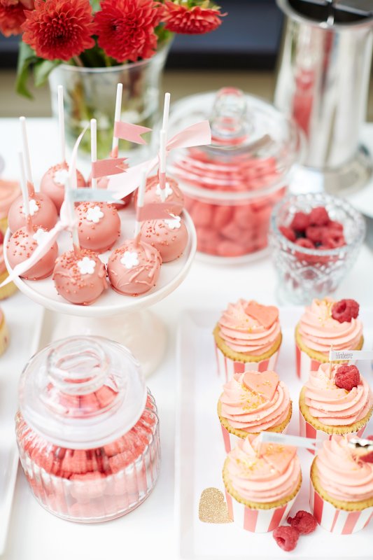 Rosa gehaltene Muffings, Cakepops und andere Leckereien auf einem Tisch mit roten Blumen im Hintergrund