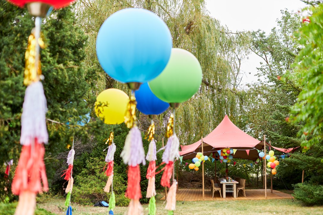 Bunt geschmückte Tafel mit Holzmöbeln und Zelt mit vielen bunten Luftballons im Freien aus der Ferne mit Luftballons im Vordergrund