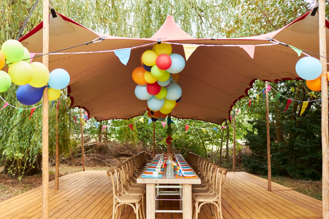 Bunt geschmückte Tafel mit Holzmöbeln und Zelt mit vielen bunten Luftballons im Freien