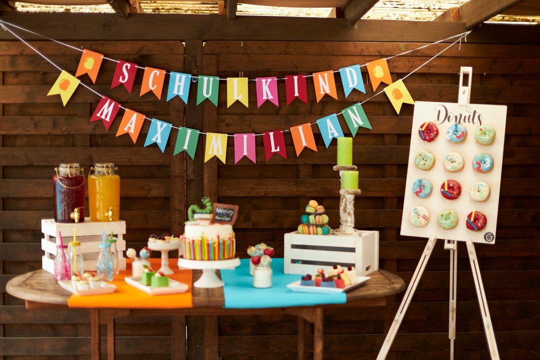 Bunt dekorierter Geburtstagstisch mit Torte und anderen Leckereien sowie einer Donuts-Tafel und einer Wimpelkette im Hintergrund