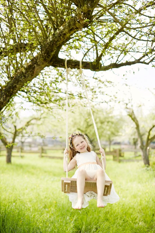 Kleines Mädchen sitzt barfuß in einem weißen Kleid mit Blumenkranz im Haar auf einer Schaukel, die an einem Baum hängt und schaukelt im Grünen