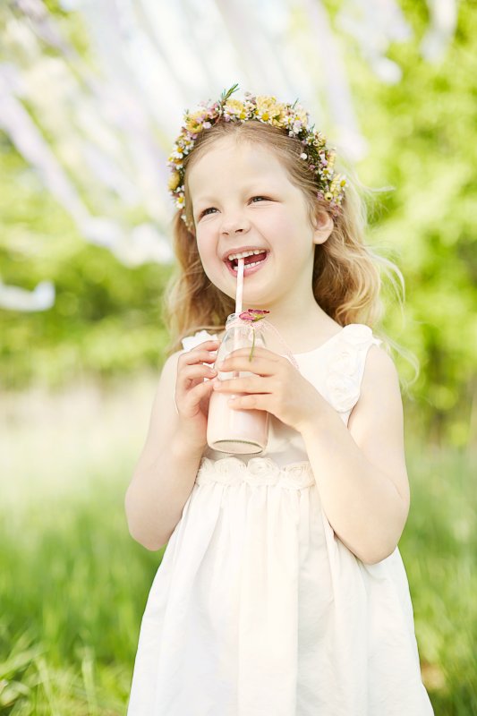 Mädchen mit weißem Kleid und Blumenkranz im Haar trinkt ein Getränk und lächelt dabei