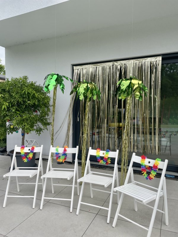 4 weiße Klappstühle mit Hawaii-Ketten vor einer Balkontür, die wiederum mit Folien-Palmen dekoriert ist