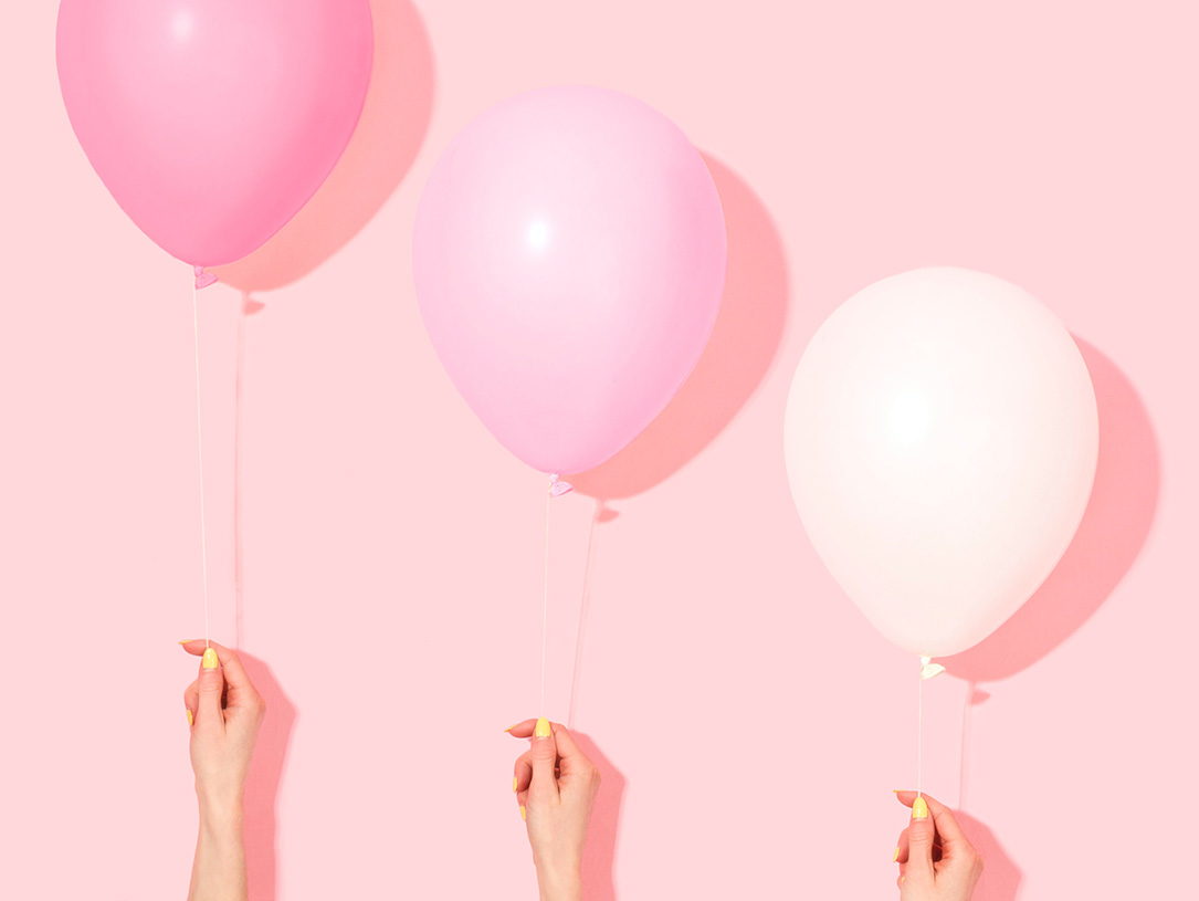 Hände halten drei Luftballons in Rosa-Tönen in unterschiedlichen Höhen vo einer rosafarbenen Wand